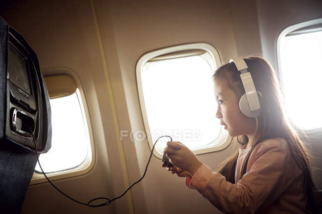 Вид збоку чарівна маленька дівчинка в навушниках грає в ігри на геймпад в літаку — стокове фото