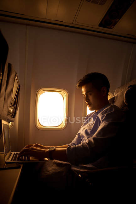 Junger Mann mit Laptop im Flugzeug sitzend, Seitenansicht — Stockfoto