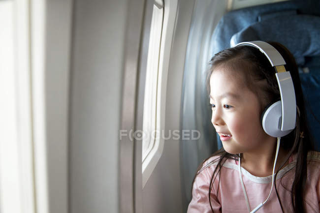 Entzückendes glückliches Kind mit Kopfhörern, das im Flugzeug sitzt und auf das Fenster schaut — Stockfoto