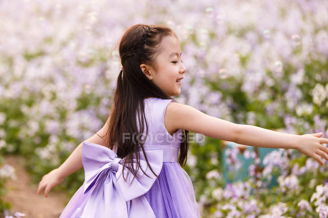 Adorable asian kid in dress catching soap bubbles at flower field — Stock Photo