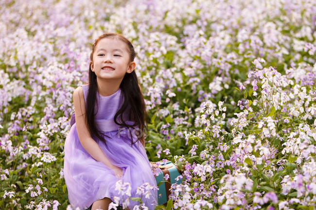 Adorable asian kid in dress sitting on bag at flower field — Stock Photo