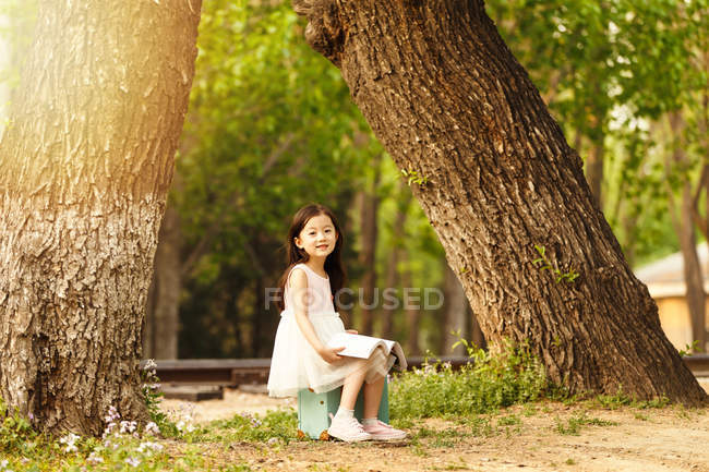 Adorable asian kid sitting on travel bag with book in park — Stock Photo