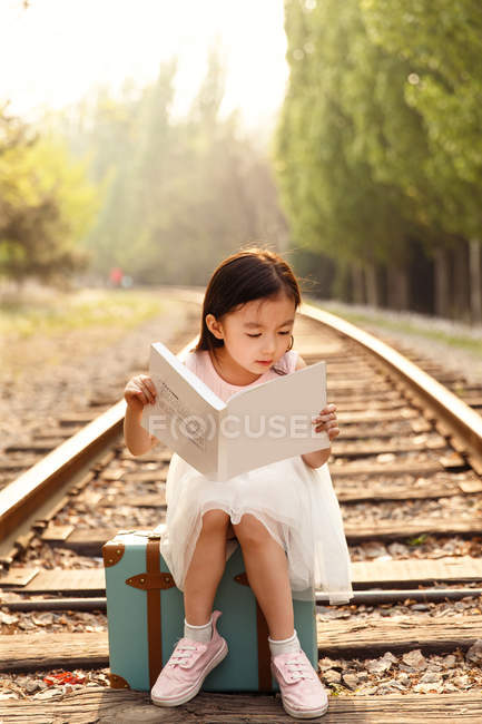 Adorable asian kid sitting on travel bag and reading book — Stock Photo