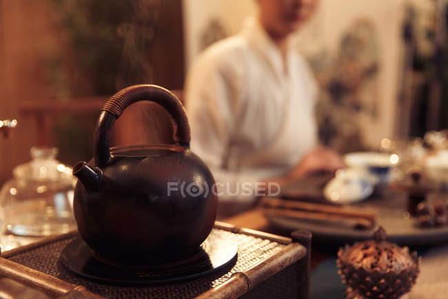 Primer plano vista de la tetera hirviendo con vapor y joven mujer asiática sentado detrás, enfoque selectivo - foto de stock