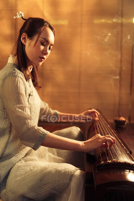 Seitenansicht einer fokussierten jungen asiatischen Frau, die ein traditionelles chinesisches Guzheng-Instrument spielt — Stockfoto