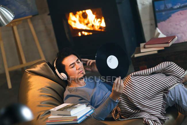 Guapo asiático hombre en auriculares celebración de vinilo registro mientras descansa en frijol bolsa silla en casa - foto de stock