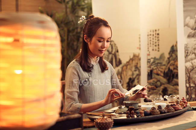 Beautiful smiling young asian woman holding dried tea leaves above table with utensils — Stock Photo