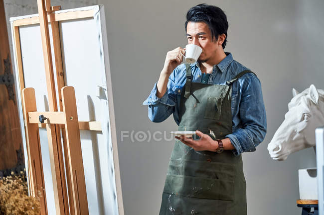 Masculino ásia pintor beber café e olhando para cavalete com foto no estúdio — Fotografia de Stock