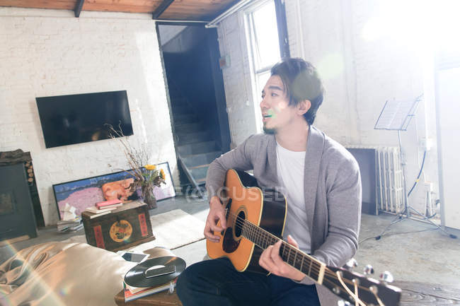 Красивий азіатський чоловік грає на акустичній гітарі і дивиться вдома — стокове фото