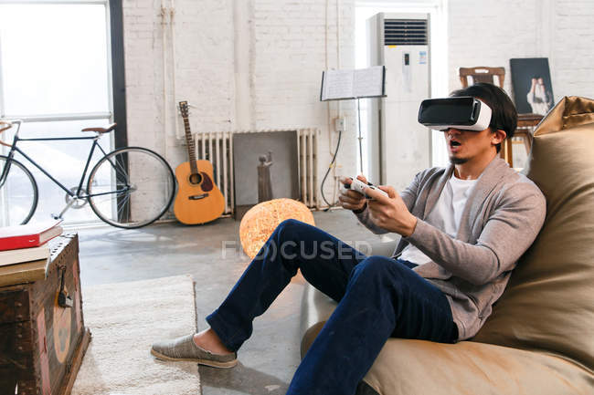 Jeune homme émotionnel en réalité virtuelle casque jouer avec joystick à la maison — Photo de stock