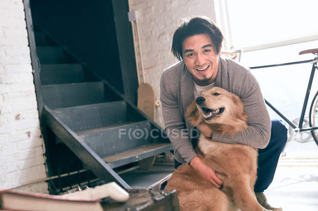 Feliz asiático homem acariciando cão e sorrindo para a câmera em casa — Fotografia de Stock