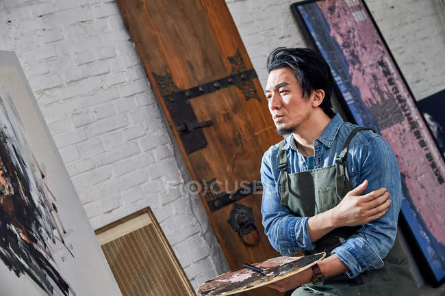 Ориентированный молодой художник в фартуке, держащий палитру и смотрящий на картину в студии — стоковое фото