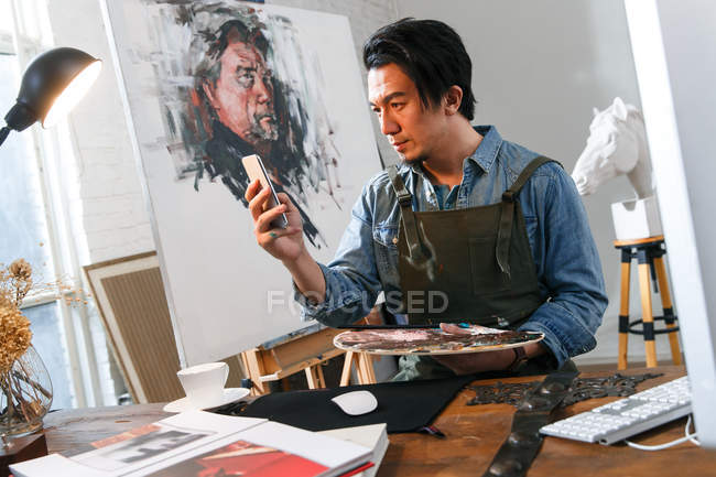 Guapo asiático pintor holding paleta y usando smartphone en estudio - foto de stock