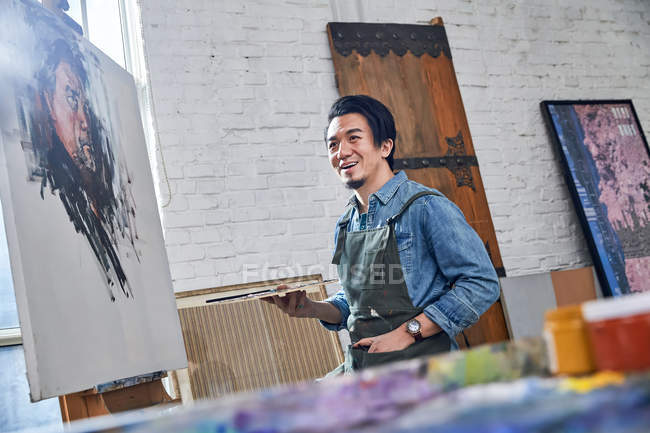 Vista basso angolo di felice giovane artista in grembiule palette holding e in piedi vicino cavalletto con ritratto in studio — Foto stock