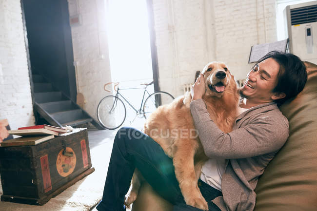 Fröhliche junge asiatische Mann sitzt auf Bohnensack Stuhl und spielt mit Hund zu Hause — Stockfoto