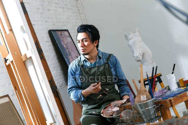 Sérieux asiatique mâle artiste tenue palette et regarder photo sur chevalet en studio — Photo de stock