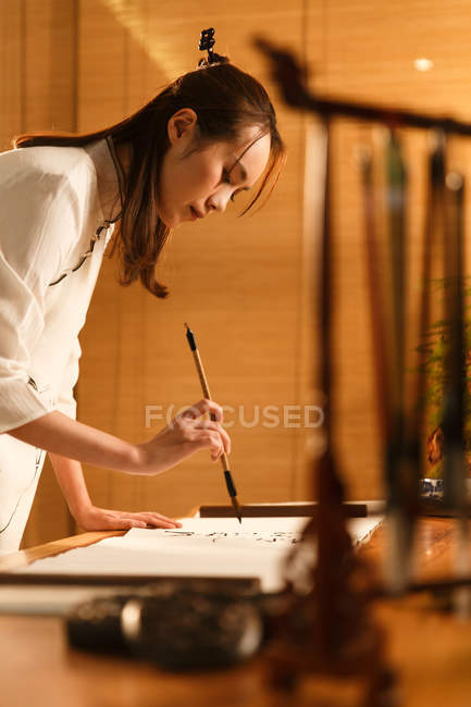 Вид збоку красива молода азіатська жінка тримає пензлик і пише китайських персонажів — стокове фото