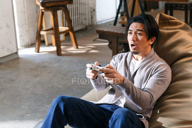 Емоційний молодий азіатський чоловік сидить на стільці з бобами і грає з джойстиком вдома — стокове фото