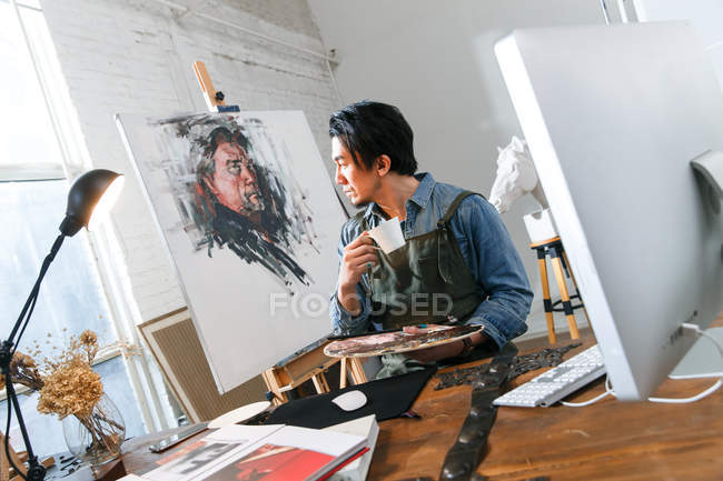 Pensive asian artist holding palette and drinking coffee in studio — Stock Photo