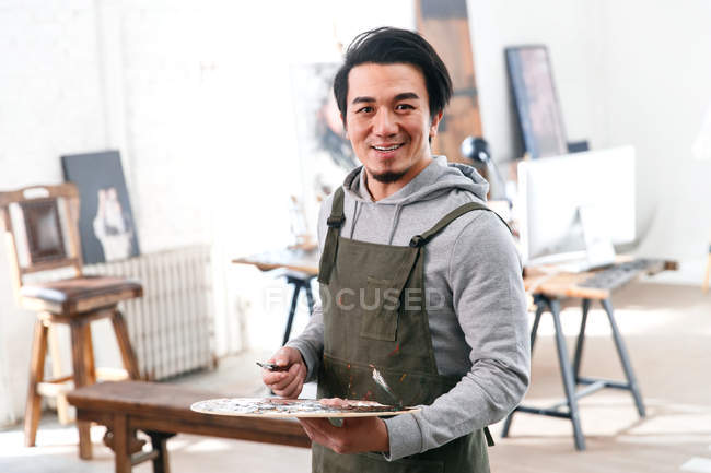 Bonito pintor masculino segurando paleta e sorrindo para a câmera no estúdio — Fotografia de Stock