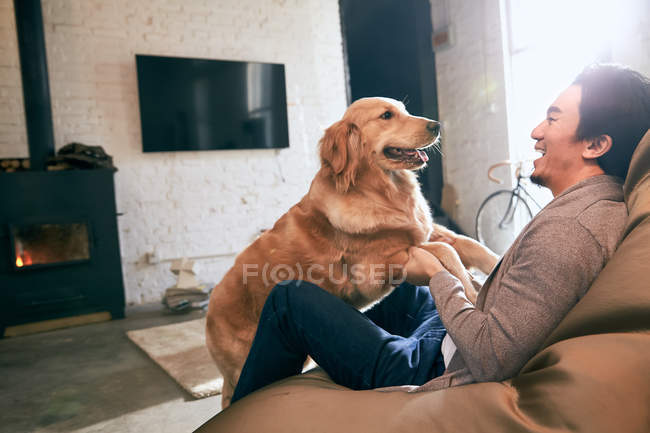 Lado vista de feliz asiático hombre sentado en frijol bolsa silla y jugando con perro en casa - foto de stock
