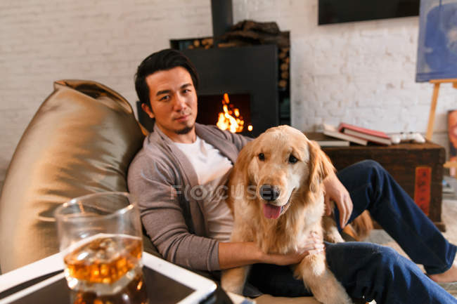 Schöner asiatischer Mann sitzt mit Hund und schaut auf Kamera zu Hause — Stockfoto
