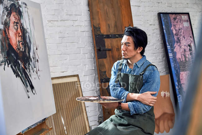 Serious male artist in apron holding palette and looking at portrait on easel in studio — Stock Photo
