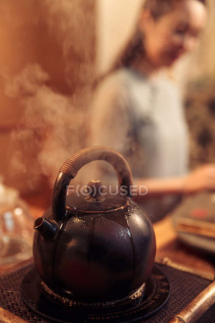 Close-up vista de bule fervente com vapor e jovem mulher asiática no fundo, foco seletivo — Fotografia de Stock
