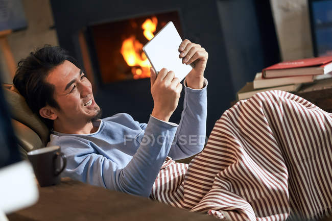 Handsome smiling asian man lying near fireplace and using digital tablet — Stock Photo