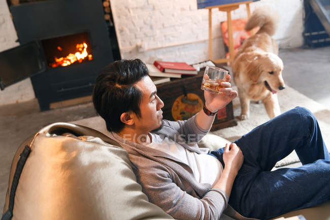 Vue grand angle de bel homme asiatique reposant dans une chaise de sac de haricot et tenant un verre de whisky à la maison — Photo de stock