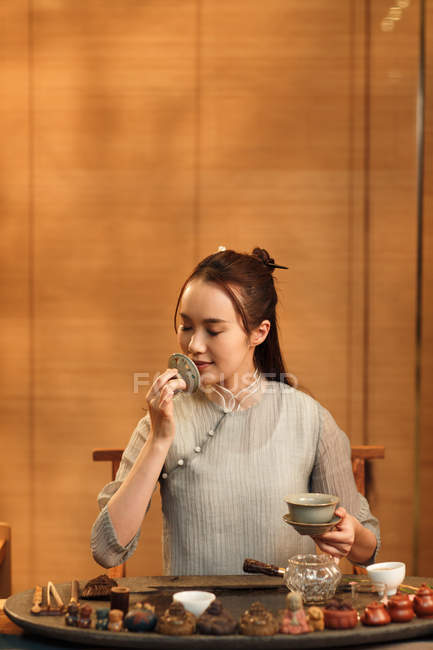 Sonriente joven mujer china sosteniendo utensilio de porcelana y oliendo té de hierbas - foto de stock