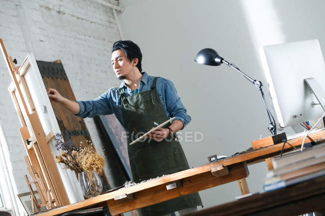 Focado artista masculino em avental segurando paleta e pintura de imagem em estúdio, visão de baixo ângulo — Fotografia de Stock