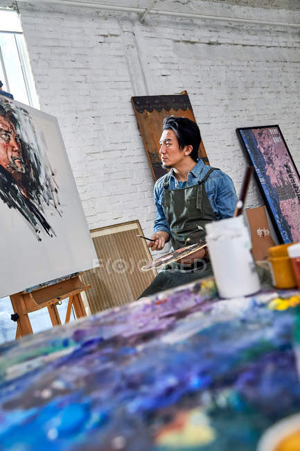 Artiste masculin concentré dans le tablier regardant le portrait en studio, foyer sélectif, outils d'art au premier plan — Photo de stock