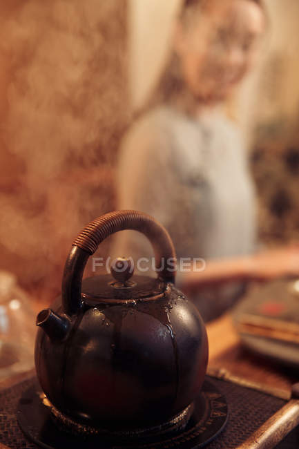 Крупный план кипящего чайника с паром и молодой азиатской женщиной сзади, избирательный фокус — стоковое фото