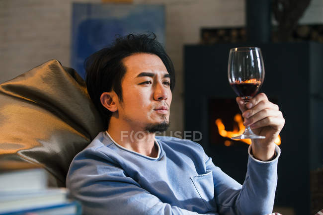 Nachdenklicher asiatischer Mann hält ein Glas Wein in der Hand und ruht sich zu Hause auf einem Bohnensackstuhl am Kamin aus — Stockfoto