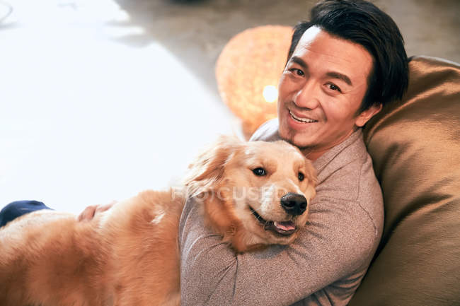 Високий кут зору веселий азіатський чоловік обіймає собаку і посміхається на камеру вдома — стокове фото