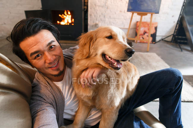 High angle view of cheerful asian man resting with dog and smiling at camera at home — Stock Photo