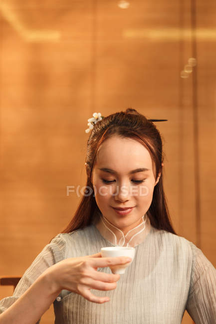 Красива усміхнена молода китайська жінка тримає чашку чаю і дивиться вниз — стокове фото