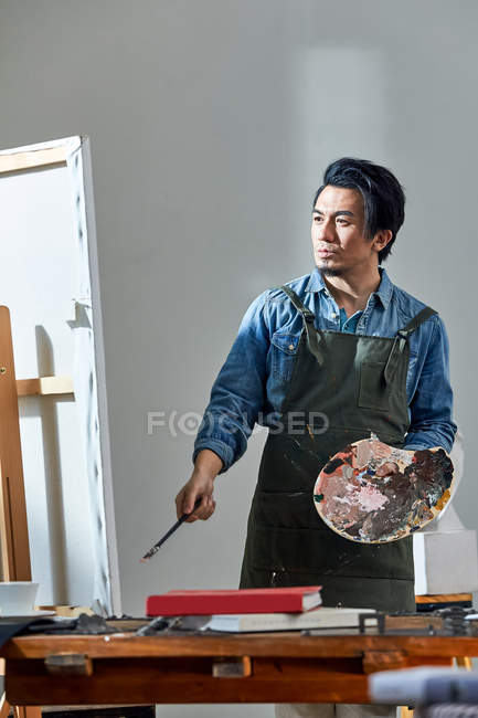 Bel artiste chinois dans tablier tenant palette et tableau de peinture en studio — Photo de stock