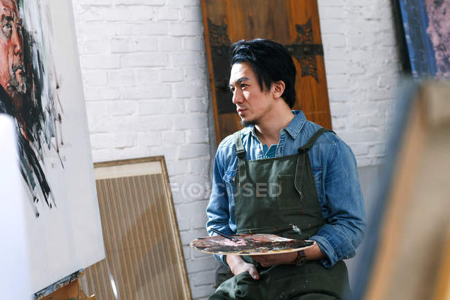 Focused young male artist in apron holding palette and looking at picture in studio — Stock Photo