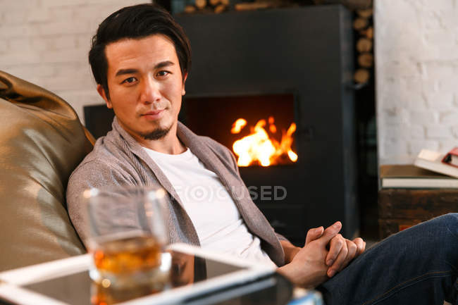 Красивий молодий Азіатська людина сидить біля каміна в домашніх умовах і дивлячись на камеру — Stock Photo
