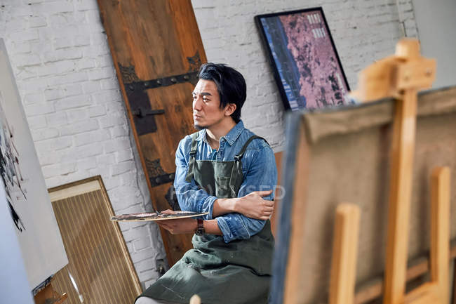 Серьезный мужчина в фартуке держит палитру и смотрит на живопись в студии — стоковое фото