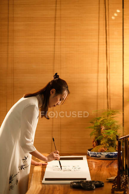 Vista laterale di bella giovane donna asiatica che tiene pennello e scrittura caratteri cinesi — Foto stock
