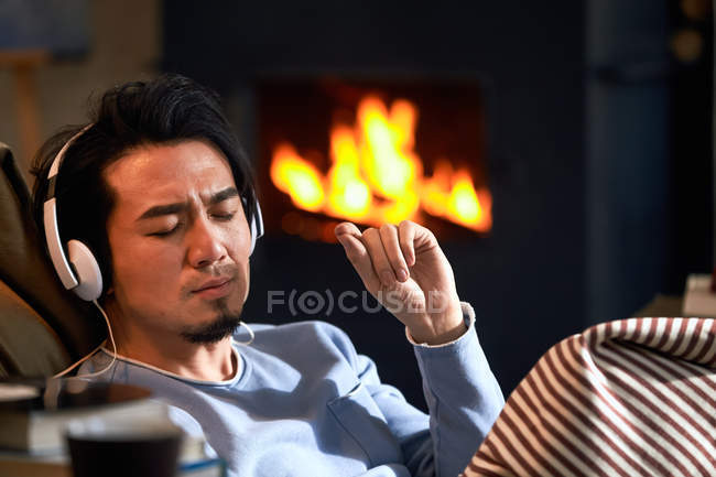 Guapo asiático hombre en auriculares escuchar música cerca de chimenea en casa - foto de stock