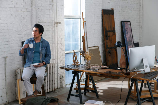 Pensive asian man holding cup of coffee and looking away in art studio — Stock Photo