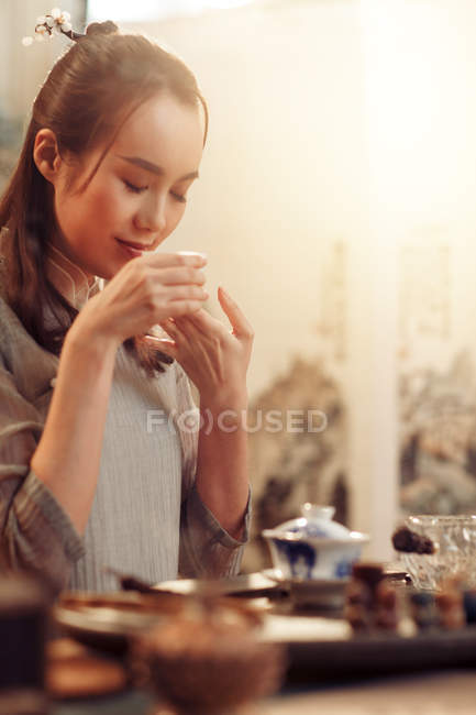 Bela sorrindo jovem asiático mulher com olhos fechados segurando copo e cheirando chá aromático — Fotografia de Stock
