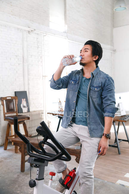Young asian man sitting on exercise bike and drinking water at home — Stock Photo
