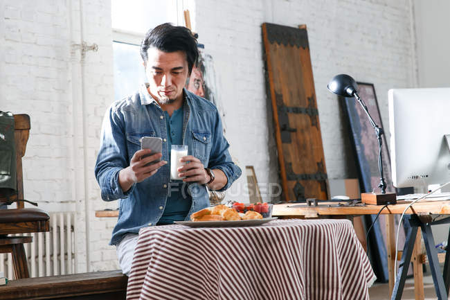 Giovane asiatico uomo bere latte e utilizzando smartphone durante la prima colazione in studio d'arte — Foto stock