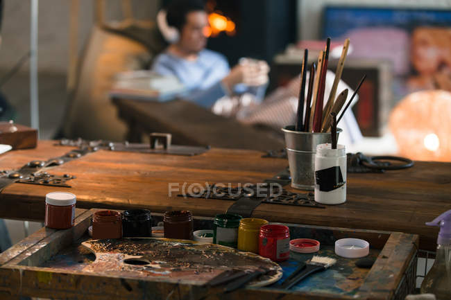 Vista de cerca de las herramientas de arte y hombre joven en los auriculares sentados cerca de la chimenea - foto de stock