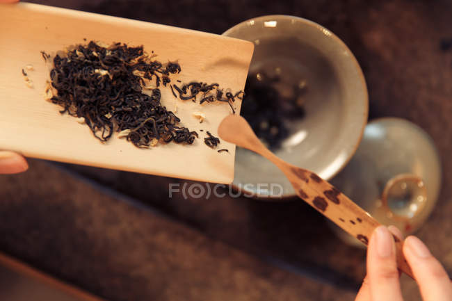 Cropped shot of woman holding wooden container with dried tea leaves above cup — Stock Photo